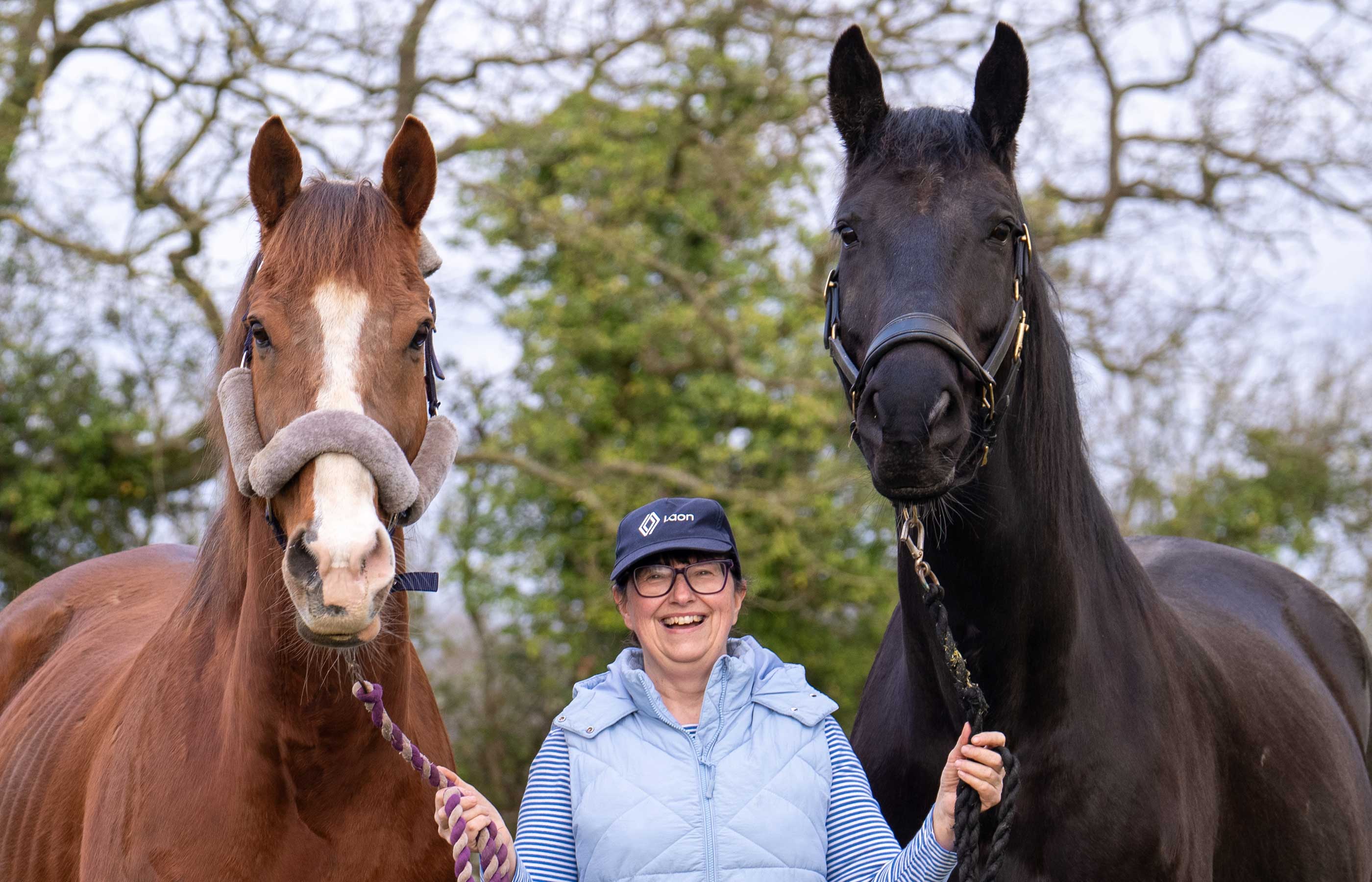 A smiling woman stands in the middle of two horses holds their reins